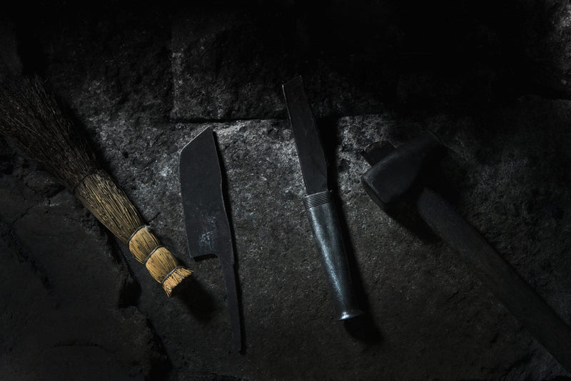 A low-key photograph of a group of tools laid on a rustic wooden table: a traditional short straw brush, two large handcrafted daggers or knives, and a metal hammer.