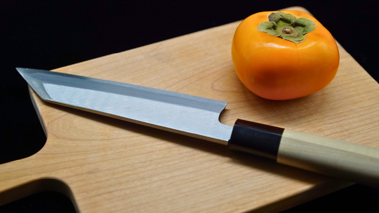 Single-bevel Japanese knife resting on a wooden cutting board next to a ripe persimmon.
