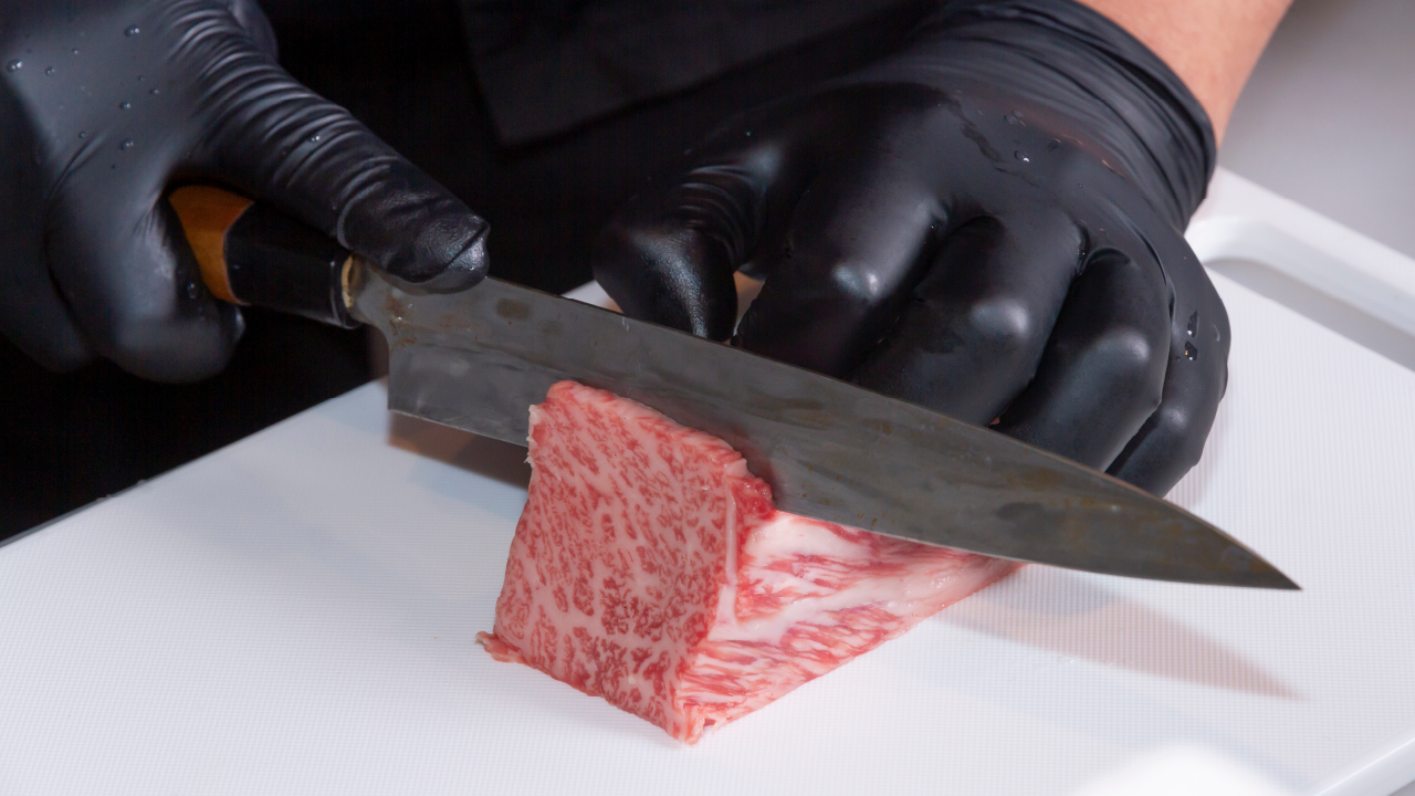 Chef wearing black gloves slicing premium marbled wagyu beef with a Japanese knife.