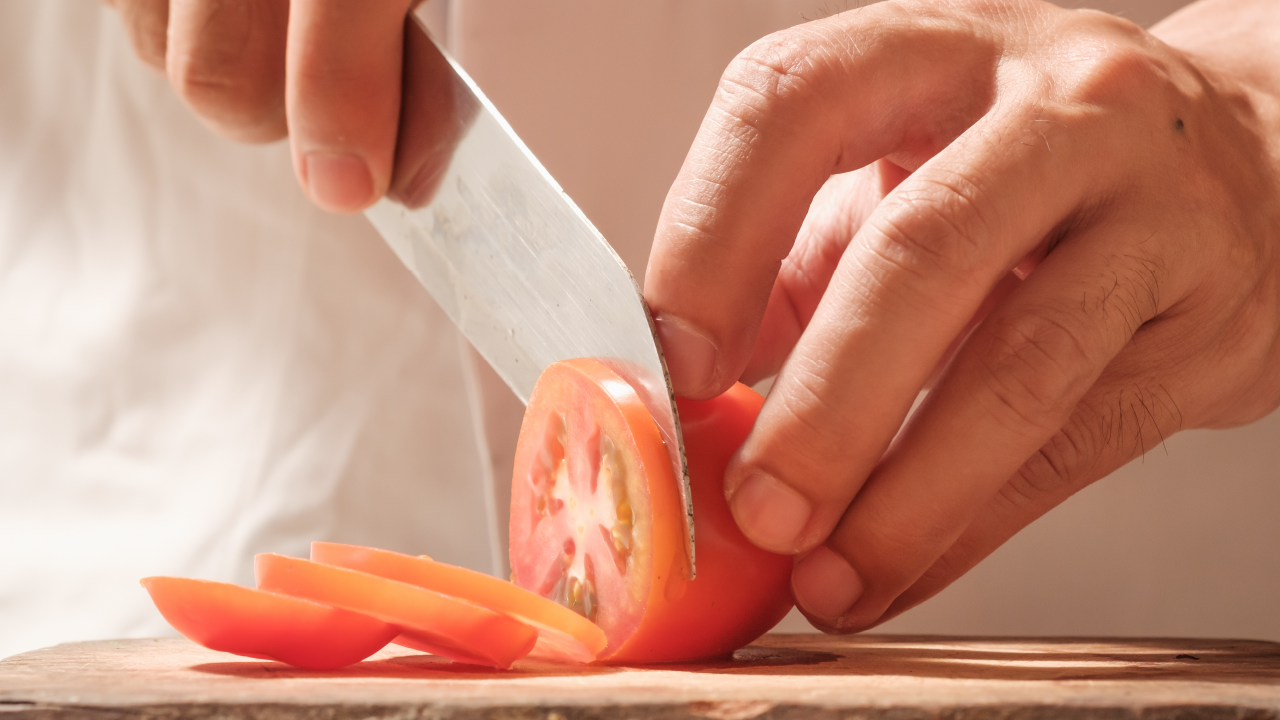 Close-up of hands slicing a fresh tomato with a sharp kitchen knife on a wooden cutting board.