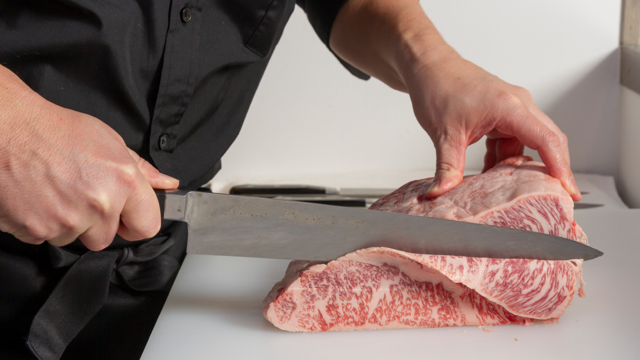 Chef cutting a large piece of marbled wagyu beef with a long Japanese kitchen knife.