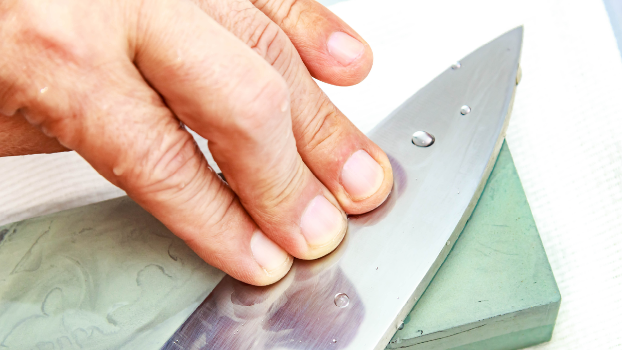 Close-up of hands sharpening a kitchen knife on a green whetstone with water droplets on the blade.