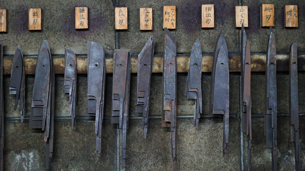Various unfinished Japanese knife blades hanging on a wall, labeled by type in wooden tags.
