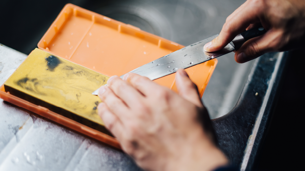 Overhead view of a person sharpening a knife on a yellow whetstone in a sink area.