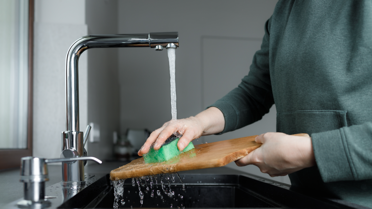 Person washing a wooden cutting board under running water using a green sponge at a kitchen sink.