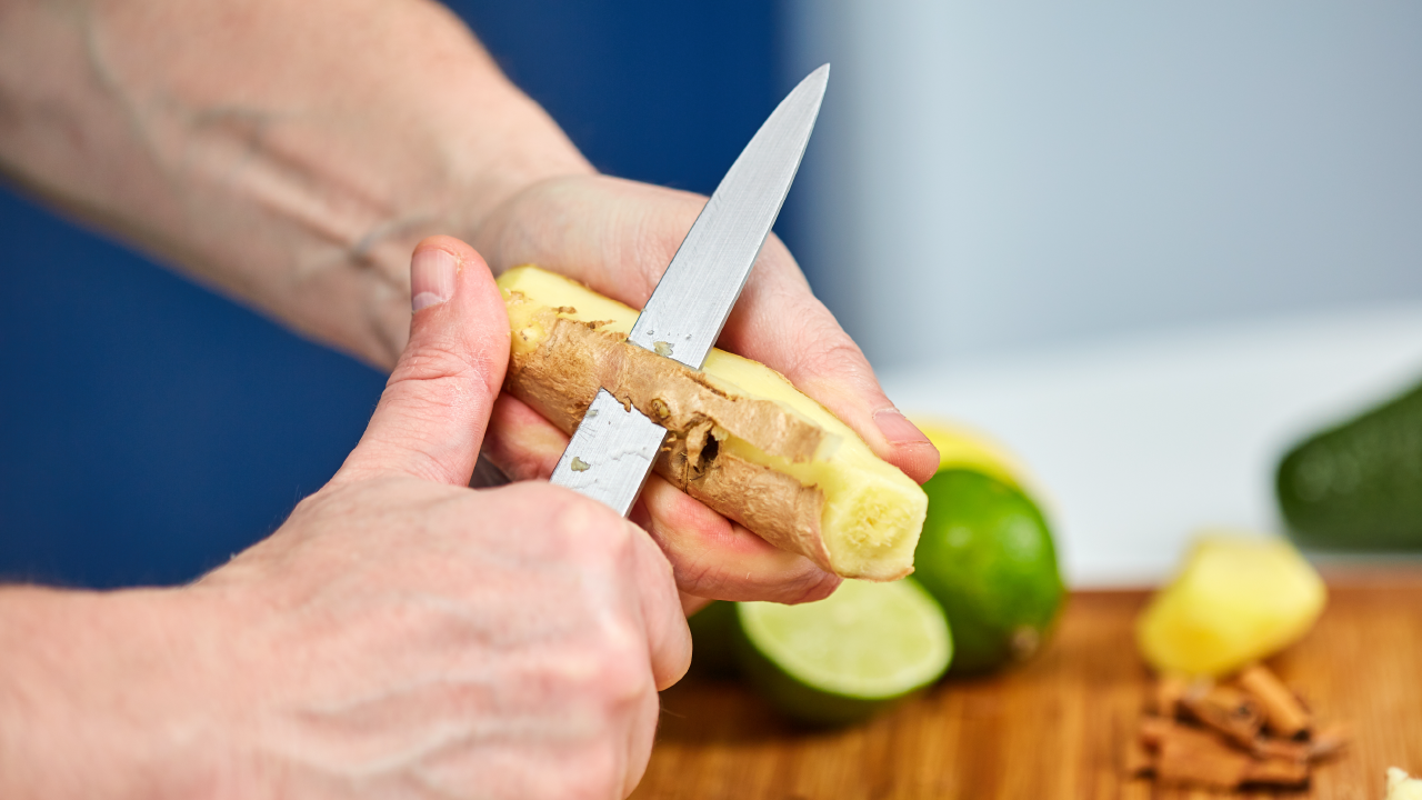 Close-up of hands peeling fresh ginger root with a small kitchen knife on a wooden cutting board, with lime and spices in the background.